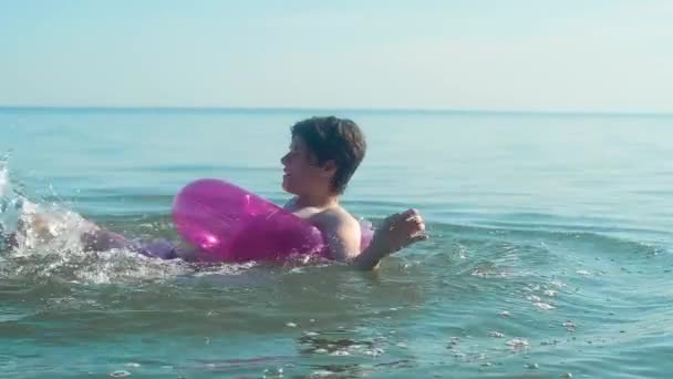 一个在海里游泳的年轻人 在粉红色的充气圆上漂浮 玩乐着用他的脚弄大水花 暑假的晴天 — 图库视频影像