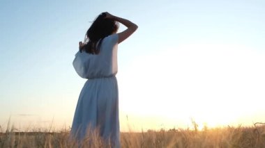 uzun koyu saçlı ve mavi bir elbise ile güzel bir genç kız dansları ve kollarını uzanmış bir buğday alanında döner. Mutlu kadın. Elleriyle saçlarını tutarak gülüyor. Doğal ışık.