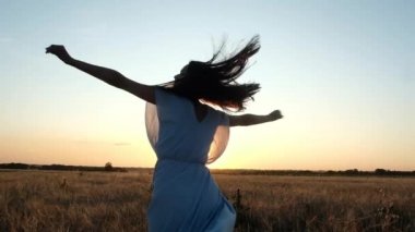 koyu saçlı güzel genç kız, mavi elbise gün batımı arka plan karşı uzanmış kollarını buğday alanında dönüyor. Mutlu kadın. Elleriyle saçlarını tutarak gülüyor. Doğal ışık.