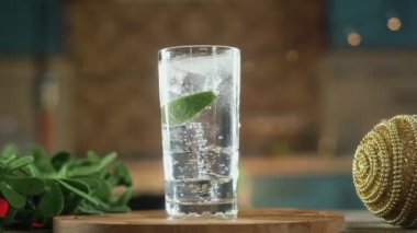 Taze sulu kireç dilimleri soda ve soğuk buz kristalleri ile cam kupa içine düşmek. Su etrafa sıçradı. Damlalar bardağa damlar. yanıp sönen Noel ışıkları karşı. Panorama çekimi