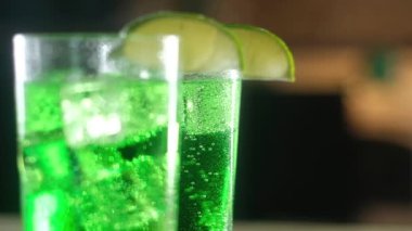 Şenlikli yeşil kokteyller. Arka planda, kenarlarında kireç parçaları olan bir bardak. Ön planda soğuk buz küplü bir bardak karbonatlı yeşil içecek var. Noel tatili