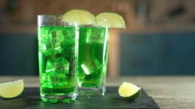 Kenarlarında limon dilimleri olan yeşil bardaklarda şenlikli kokteyller yüzer ve köpüklü içeceklerle fırıl fırıl döner. Meyveli limon dilimleri tepside içkinin yanında yatıyor. Noel tatili. Ağır çekim. Ortalama plan.