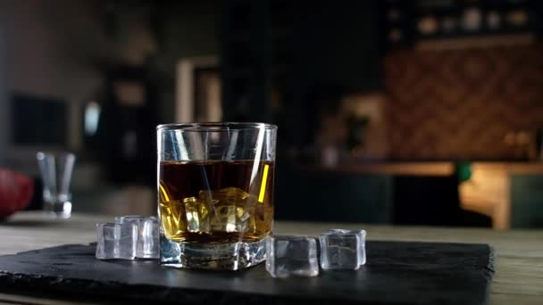 大きな氷のキューブと強いシングルモルトウイスキーの明確なガラスのパノラマショット ガラスは黒い台の上にある 氷の結晶の横に置く 愛好家のためのお祭りアルコール飲料 — ストック動画