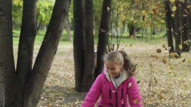 Pembe ceketli ve uzun saçlı kot pantolonlu genç kız bir yığın sarı yaprak fırlatıyor. Sonbaharda bir çocuk parkta eğleniyor. Yaprakları fırlatır ve neşeyle güler. yavaş çekim