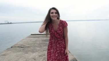 Kırmızı elbiseli, siyah şapkalı, uzun saçlı, beyaz bir kadın rıhtımda kameraya gülüp flört ediyor. Kız neşeli bir şekilde gülüyor..