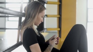 Çekici, zayıf atletik kız ringin yanında, halatların yanında, kum torbalarının arka planında oturuyor. Bir kadın spor yaptıktan sonra kalori saymak için akıllı telefonunda uygulama kullanıyor..