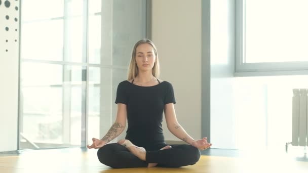 女性は大きな窓の前のトレーニングルームでロータスのポジションで瞑想します 痩せっぽちの運動女子が床に座って息をする 集中運動をしています 健康的なライフスタイルの概念は — ストック動画