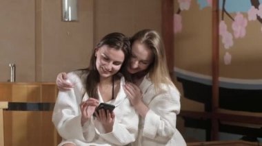 Kafalarında havlu olan beyaz önlüklü güzel seksi kadınlar kaplıca salonunda oturuyorlar. Kızlar prosedürlerden sonra rahatlayın ve telefonu eğlence için kullanın. Yakın çekim bakım ve cilt bakımı.