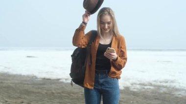 Sarı saçlı, sırt çantalı güzel bir kız sahilde şapka takıyor. Kadın modern akıllı telefon kullanıyor ve mesajları çevirirken gülüyor. Seyahat ederken doğada özgürlük ve rahatlama