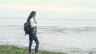 Sırtında sırt çantasıyla genç bir kız deniz uçurumunun kenarında duruyor. Tatillerde ya da hafta sonlarında bavulla denize git. Sırtında sırt çantasıyla gezen bir kız