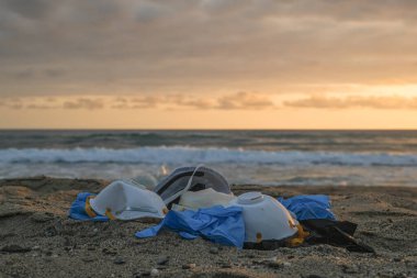 Koruyucu virüs maskesi ve plastik eldiven. Günbatımı deniz kıyısında çöplük. Coronavirus covid kirliliği. 