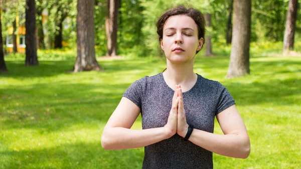 Young calm woman practicing yoga,relaxing meditating namaste gesture,exercising in Padmasana.Mindful peaceful caucasian girl in grey t-shirt practicing breathing yoga in green park outdoor.No stress