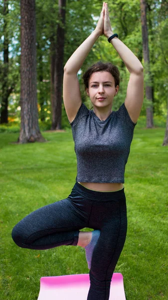 Smiling young woman practicing yoga in green city park outdoor.Cute girl in grey sportswear work out standing in Vrksasana exercising with namaste gesture overhead,tree pose.Healthy lifestyle.Vertical