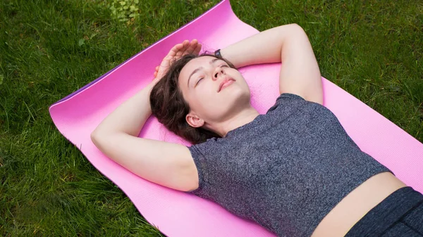 Gri bluzlu genç kadın gözlerini shavasana 'ya kapamış yatıyor, yoga sonrası ceset duruşu, egzersiz egzersizleri, antrenmandan sonra dinlenme, meditasyon, yeşil çimlerin üzerinde derin derin nefes alma.