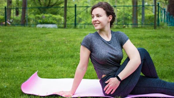 Yoga esneme egzersizi, şehir parkında yeşil çimenlere bel altı geren mutlu, beyaz bir genç kadına uyuyor. Spor paspasının üzerinde oturan sağlıklı bir kız. Açık havada oturmuş omurga bükülmesi.