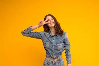 Mutlu, sevimli, iyimser, kıvırcık, esmer, kot elbiseli, dişlek, gülümseyen, barışçıl zafer jestini gösteren, disko işareti taşıyan, sarı başlıklı kadın.