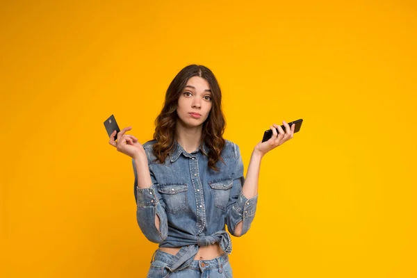 Upset confused young woman shrugging with smartphone and credit card in hands,looking at camera isolated on yellow background.Debt problems,insecure online payment,failed transaction,money