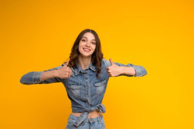 Mutlu genç bir kadın işaret veriyor ve neşeyle gülümsüyor, birine destek ve saygı gösteriyor. Esmer kız sarı arka planda izole edilmiş gibi el kol hareketi yapıyor. Vücut dili..