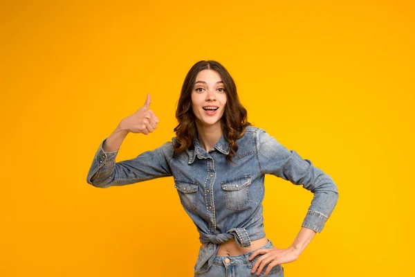 Good job.Emotional smiling curly brunette woman wearing denim clothes show thumb up approval like gesture,support satisfied recommend the best.Standing isolated on yellow background.