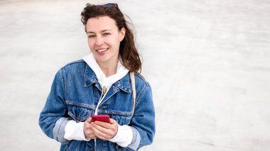 Havalı kot ceketli, elinde cep telefonu olan, kameraya sevinçle gülümseyen beyaz bir kadın. Hippi kız açık havada beton arka planda duruyor, haberleri güncelliyor, haberleri paylaşıyor.