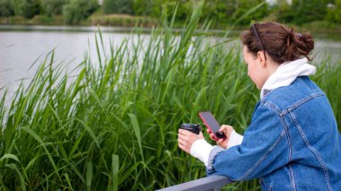 Mavi kot ceketli, elinde cep telefonu ekranına bakan, elinde göl kenarındaki raylara yaslanmış kahve fincanı tutan esmer bir kadının görüntüsü. Sohbet, gezinme, blog yazma, online alışveriş, kaydırma.