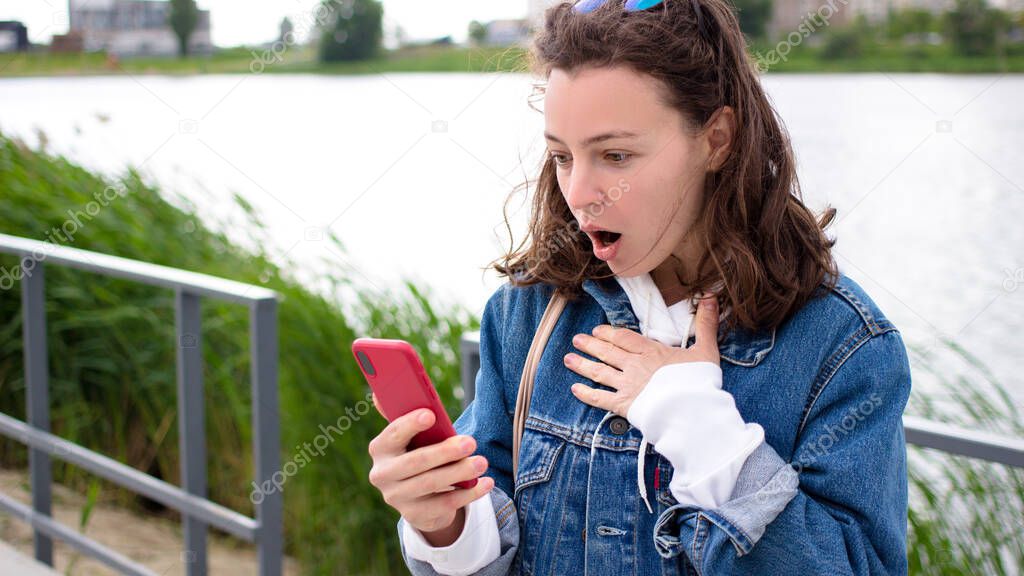 Surprised amazed girl looking at mobile phone screen with open mouth standing outdoor near city lake.Astonished teenager hipster student customer shocked by social media online content hold cellphone