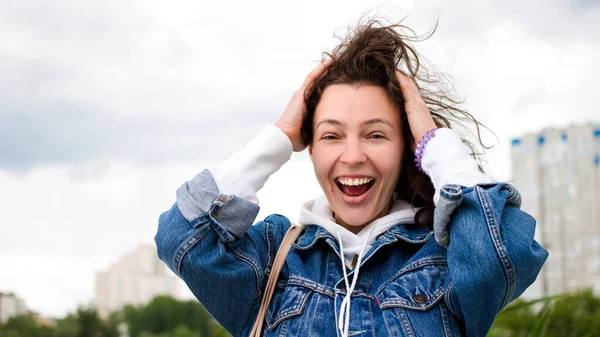 Happy emotional excited brunette caucasian girl holding head being amazed,standing outdoor city buildings and green park background.Teenager hipster student smiling surprised,shocked after good news