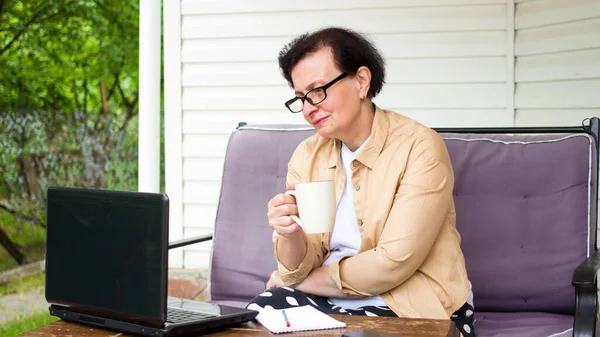 나이든 여성이 노트북 컴퓨터 화면을 커피를 마시는 테라스에 정원에서 뉴스를 — 스톡 사진