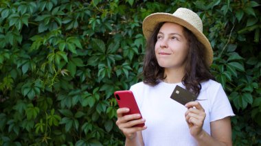 Banka kartı, cep telefonu tutan genç bir kadın, sevinçle yukarı bakıyor, yeni elbise elbiselerini düşünüyor. Yaz şapkalı bir kız internet ödemesi yapıyor, açık havada online alışveriş yapıyor. 