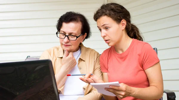 Serious senior woman and grown up daughter looking at laptop screen,calculating family budget,planning expensive purchase,discussing finance.Young woman helps mature mother pay domestic debts online.