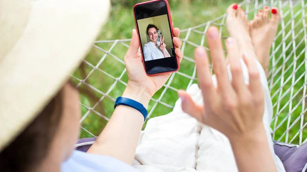 Phone video call.Young woman talking with her friend by video call,gesturing hi to her.Girl making video conference with smartphone relaxing lying in hammock in green garden outdoors.Selective focus.