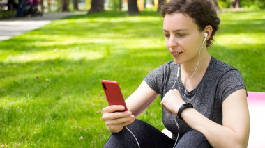Kulaklıklı, akıllı telefonlu genç bir kadın, ekrana bakıyor, çevrimiçi antrenman izliyor, egzersiz sonrası sosyal medyada aktivite verilerini paylaşıyor, fitness minderinde oturuyor, şehir parkında dinleniyor.