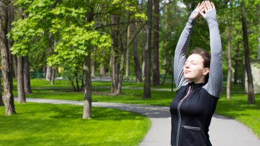 Formda ve sportif genç bir kadın yeşil şehir parkında esneme hareketleri yapıyor. Esmer, mutlu, gülümseyen, atletik bir kız. Eğitimden önce el kaldırıyor, sabah egzersizi, spor yapıyor, güneşli bir günde koşuyor.