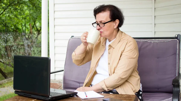 노인 여성이 커피를 마시고 노트북 컴퓨터 화면을 보고, 집 앞 테라스에 앉아 있습니다. 조용 한 브루 네트 성숙 한 여성, 정원에서 편안하게 잔을 들고, 뉴스를 보고, 온라인 웹 사이트, 비디오 — 스톡 사진