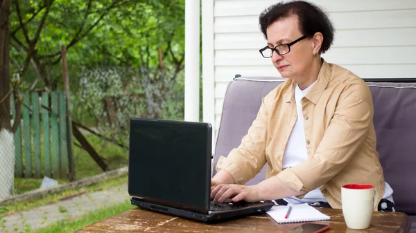 Serious focused senior woman professional employee typing on laptop computer,sitting at home terrace office table, mature student studying e learning online course,using technology app for work