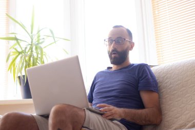 Milenyum sakallı, gözlüklü, dizüstü bilgisayara hayret içinde bakan, evdeki koltukta oturan bir adam. İnanılmaz haberler okuyan, çevrimiçi dükkanda kazançlı bir teklifle şaşıran, video film izleyen.