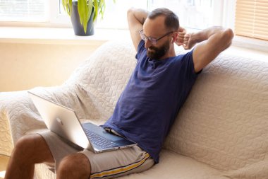 Gözlüklü mutlu genç adam, kollarını uzatan modern kulaklıklar, koltukta oturuyor, evde online işten sonra dizüstü bilgisayara bakıyor. Günün sonunun keyfini çıkarıyorum, projenin başarıyla bittiğini hissediyorum..