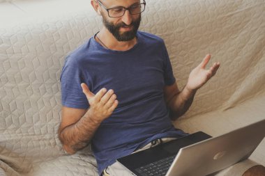Bin yıllık gözlüklü, kablosuz kulaklık takan, müşterilerle video görüşmesi yapan, evdeki kanepede oturan bir iş adamı. Çevrimiçi konferans, webinar, eğitim sınıfı dersi.