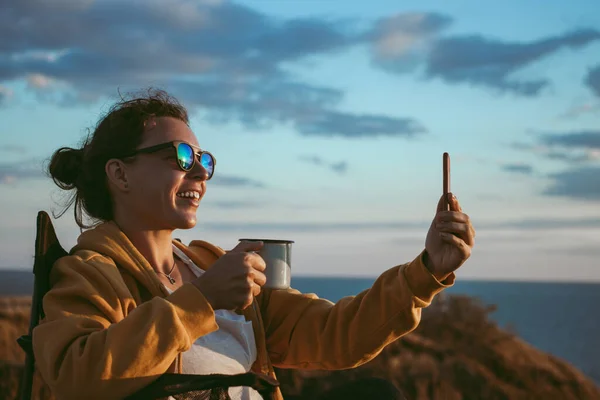 Happy young woman making video call with smartphone, sharing data on social media, holding iron mug cup with coffee, sitting in touristic chair on cliff in front of sea on sunset. Technology concept.