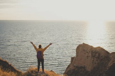 Sırt çantalı, dağ zirvesinde kollarını açmış, gün batımında kayanın üzerinde durup denize bakan mutlu genç kadın yürüyüşçünün arka görüntüsü. Güçlü ve özgürlük yaşam tarzı konsepti. Macera gezisi. 