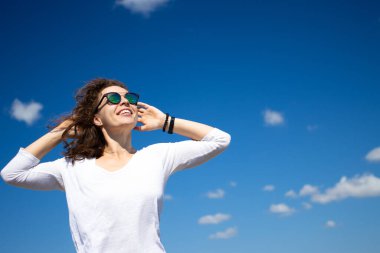 Tasasız, güzel, esmer, yüzü ve elleri gökyüzünde yükselmiş, temiz hava soluyan güneşli günün tadını çıkaran, rüzgarı estiren, mavi arka planda duran genç bir kadın. Özgürlük, stres yok yaşam tarzı konsepti