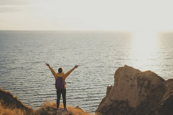 Sırt çantalı, dağ zirvesinde kollarını açmış, gün batımında kayanın üzerinde durup denize bakan mutlu genç kadın yürüyüşçünün arka görüntüsü. Güçlü ve özgürlük yaşam tarzı konsepti. Macera gezisi. 