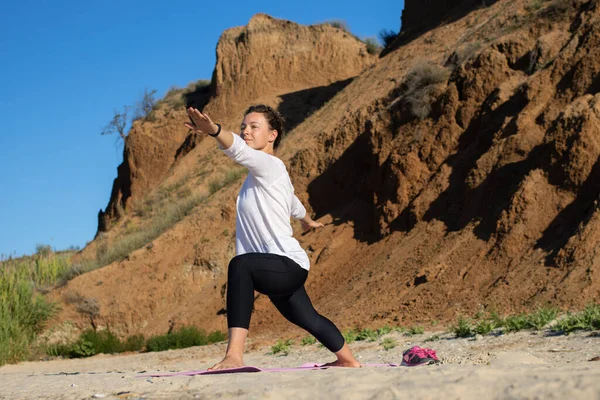 Genç esmer kadın sabahları kumsalda yoga yapıyor, savaşçı pozunda duruyor, Virabhadrasana kumlu kayalıkların ve mavi gökyüzünün arka planında. Ruh sağlığı ve öz bakım kavramı. 