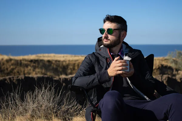 Güneş gözlüklü ve su geçirmez ceketli genç esmer adam elinde demir bardak kahve, deniz kenarındaki bir kamp sandalyesinde tek başına oturuyor, doğa manzarasında tatilin tadını çıkarıyor. Seyahat, aktif yaşam tarzı.