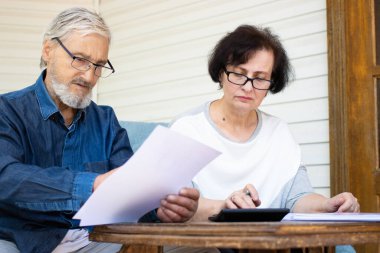 Ciddi odaklı yetişkin çift kağıt belgeler okuyor ve iç finans, aile yönetimi, planlama bütçesi, harcamalar, ev teraslarındaki kanepede oturarak ödeme veya kontrol faturaları hesaplıyor.