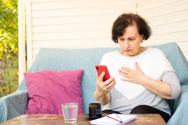 Nervous frustrated brunette senior woman holding mobile phone and looking at screen while reading disturbing news in media, watching shocked video content, sitting on sofa on home terrace outdoors — Stock Photo, Image