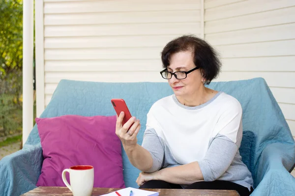 Güzel, orta yaşlı, gözlüklü esmer kadın cep telefonu ekranına bakarken mesajları kontrol ediyor, haberleri okuyor, kablosuz internet bağlantısı kullanıyor, evdeki terasta kanepede oturuyor.