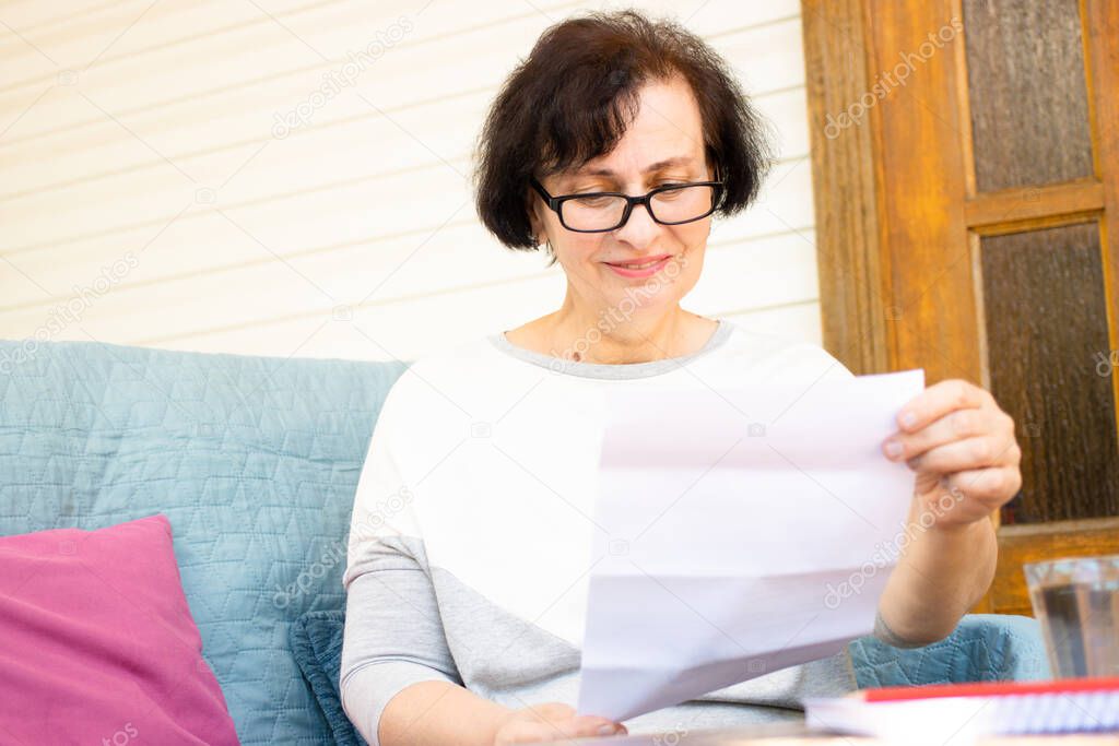 Happy middle aged elderly woman in eyeglasses reading paper document or postal letter from friend, sitting on sofa on home terrace. Rejection of pleasant paperwork bank notice or post correspondence