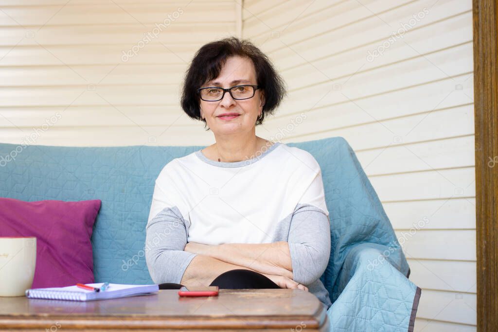 Head shot portrait of happy brunette middle aged senior woman in eyeglasses, smiling pleasant looking at camera sitting on sofa on home terrace, enjoying free leisure weekend time alone