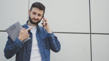 Yakışıklı sakallı genç adam akıllı telefondan konuşuyor gülümsüyor, eli ve parmağı ile işaret ediyor, kot gömlek giyiyor, şehir duvarının arka planında dikiliyor. Boşluğu kopyala.
